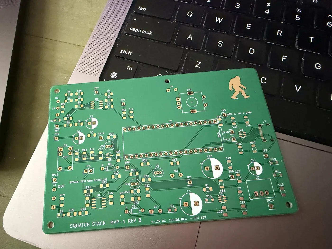 A bare green circuit board, silkscreened “Squatch Stack MVP-1 rev B” with the walking Squatch in gold, resting on a laptop keyboard